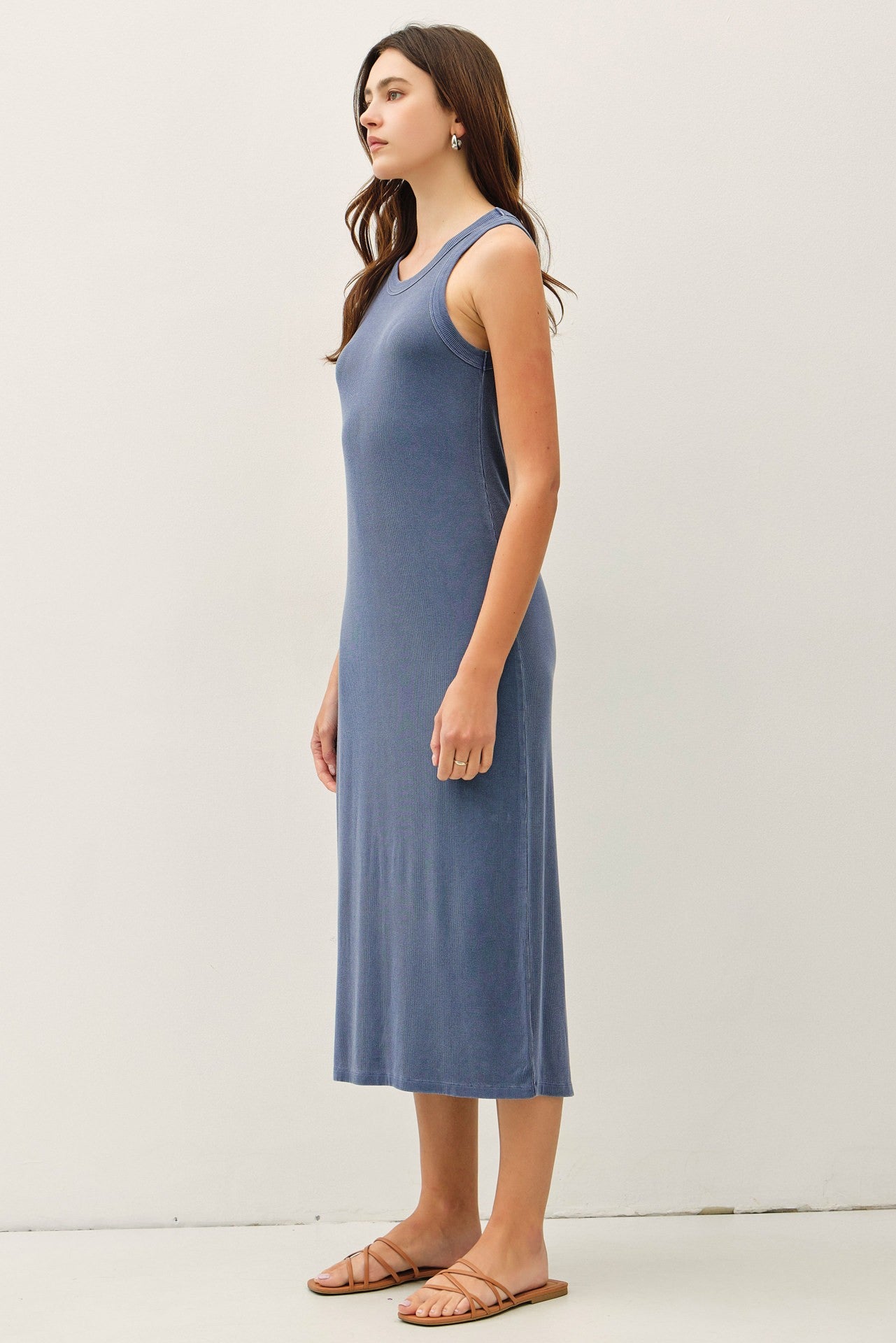 woman wearing a sleeveless blue ribbed midi dress and brown sandals, standing sideways against a plain background