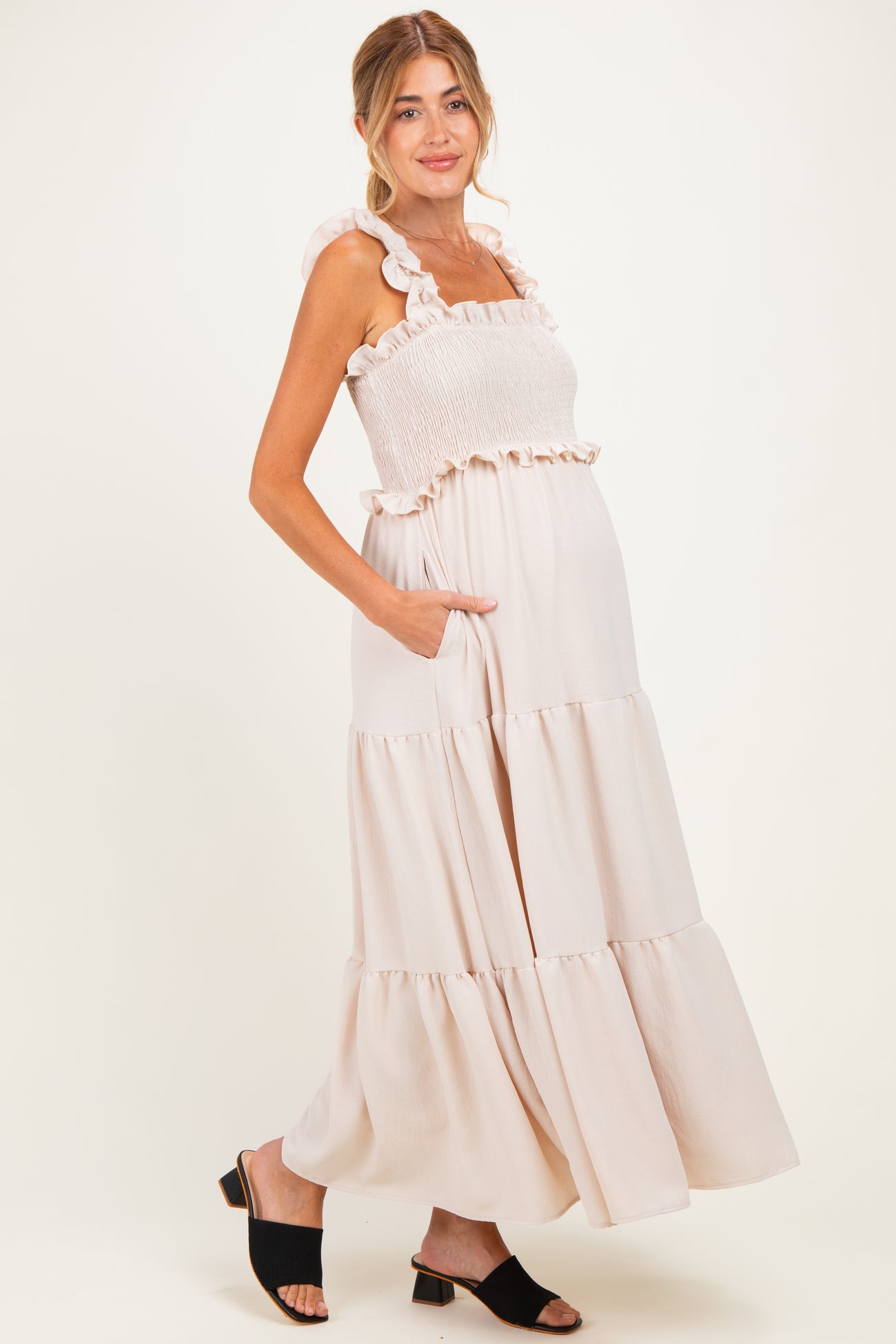 woman wearing a light beige maternity maxi dress with ruffle details and pockets, showing her baby bump