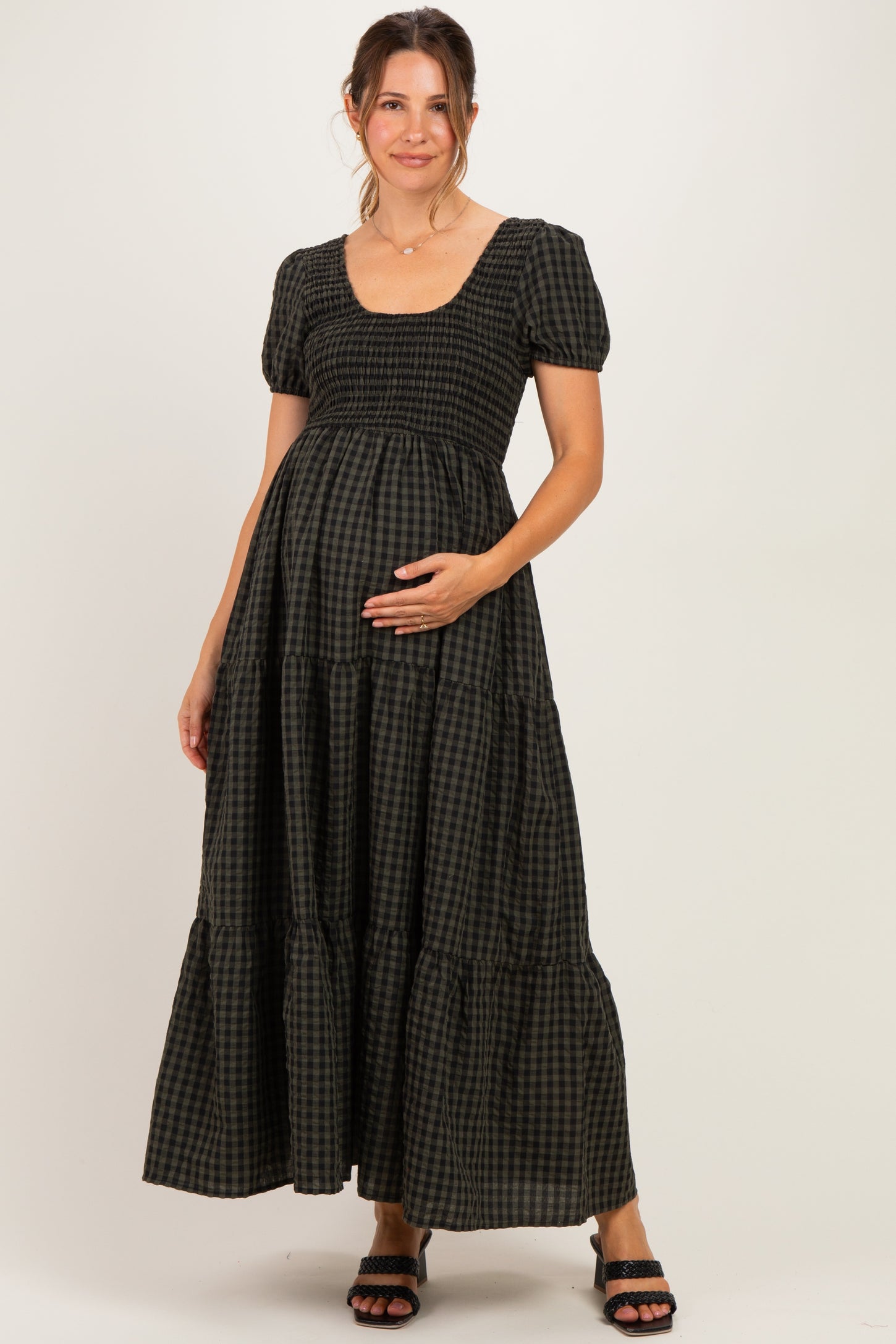 woman wearing a black and green checked maternity dress with short puff sleeves and a tiered skirt, cradling her baby bump