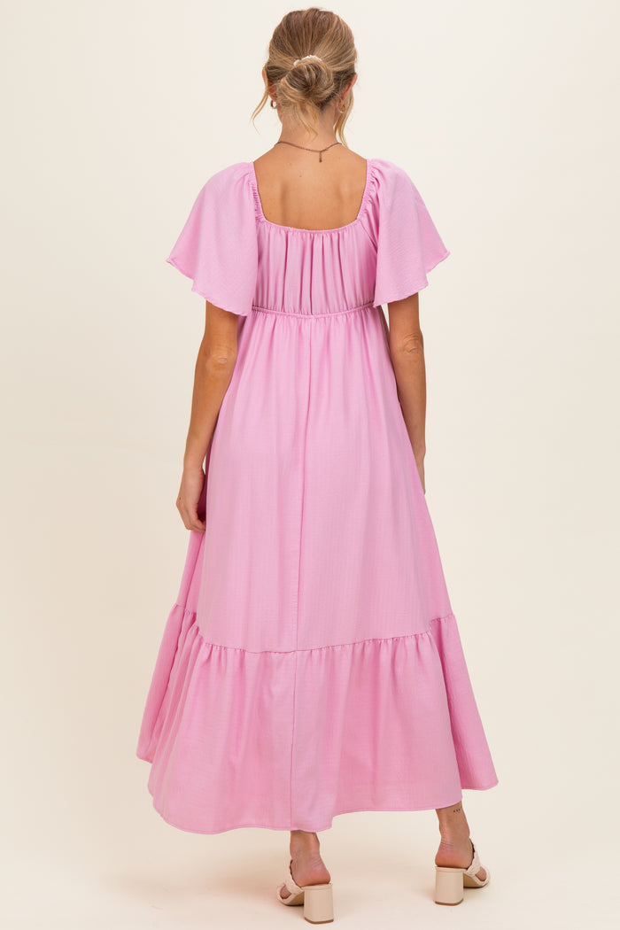 woman wearing a light pink maternity maxi dress with flutter sleeves and a tiered skirt, showing her baby bump from the back