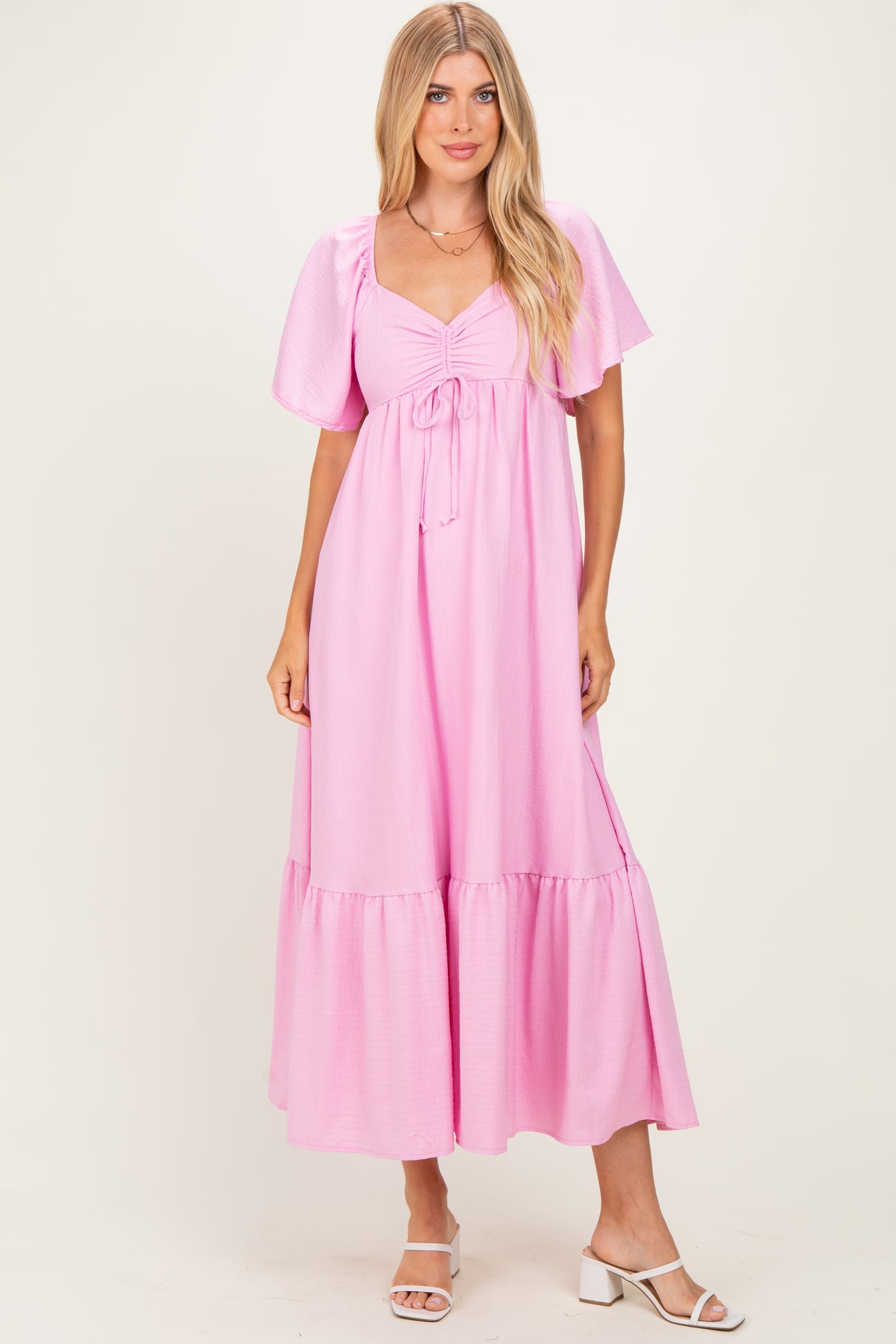 woman wearing a pink maxi dress with puff sleeves and a tie detail at the bust, standing in a light background.
