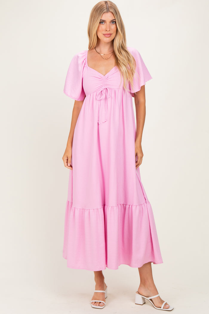 woman wearing a pink maxi dress with puff sleeves and a tie detail at the bust, standing in a light background.