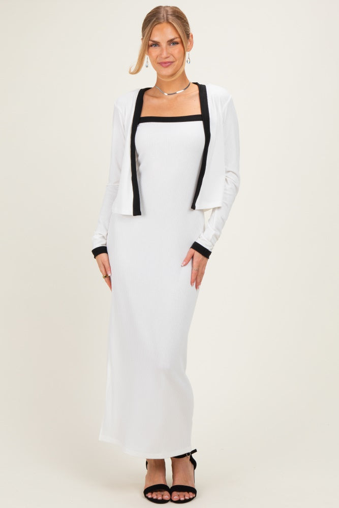 woman wearing a white maxi dress with black trim and a matching cropped jacket, posing with one hand on her hip