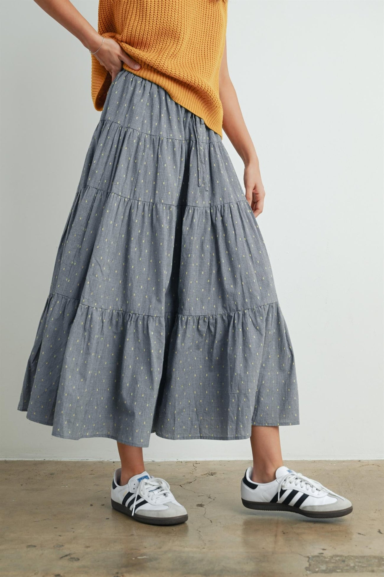 woman wearing a gray tiered midi skirt with yellow polka dots and white sneakers, paired with a relaxed orange sweater