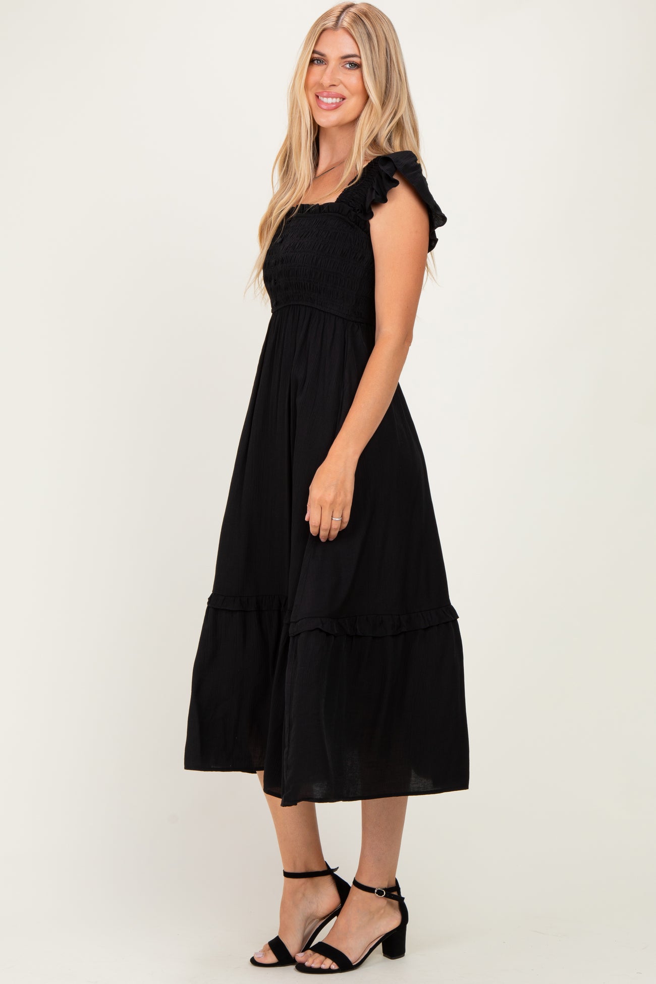 woman wearing a black smocked midi dress with ruffle straps and a tiered skirt, standing and smiling