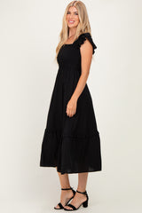 woman wearing a black smocked midi dress with ruffle straps and a tiered skirt, standing and smiling