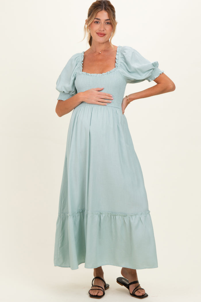 woman wearing a light blue maternity dress with puff sleeves and a tiered skirt, gently holding her baby bump