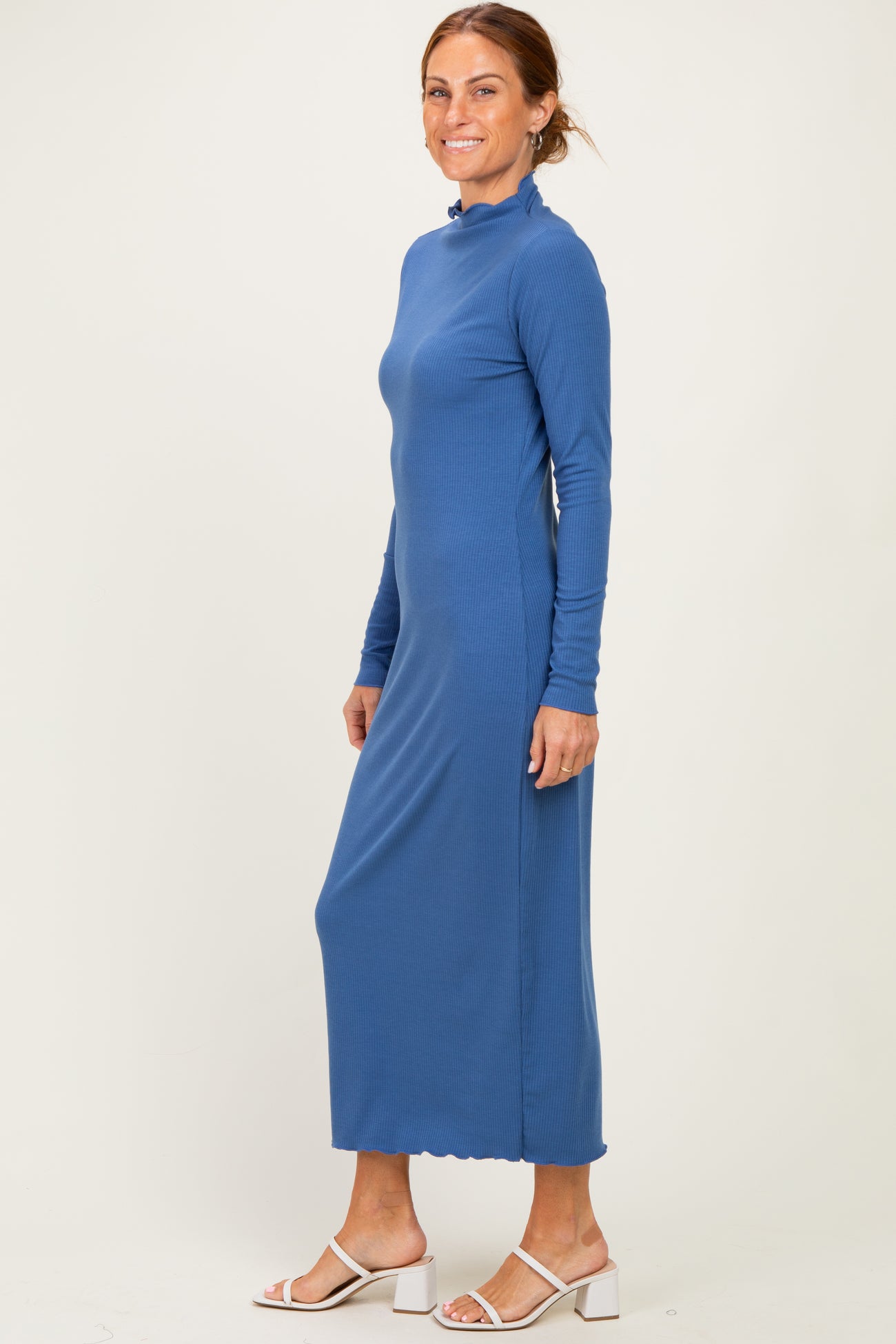 woman wearing a long sleeve blue ribbed maxi dress with a high neck and scalloped hem, standing sideways against a light backdrop
