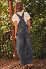 woman wearing a pair of denim overalls with a white t-shirt underneath, standing against a leafy background