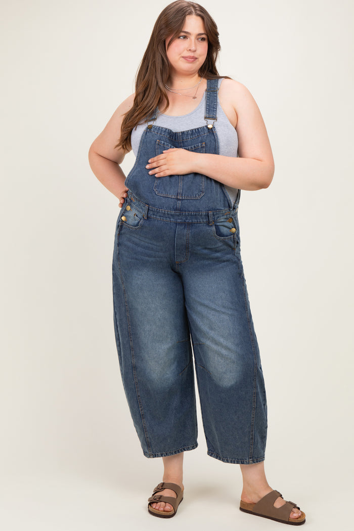 woman wearing a blue denim maternity plus overalls with a gray tank top and standing with one hand on her hip