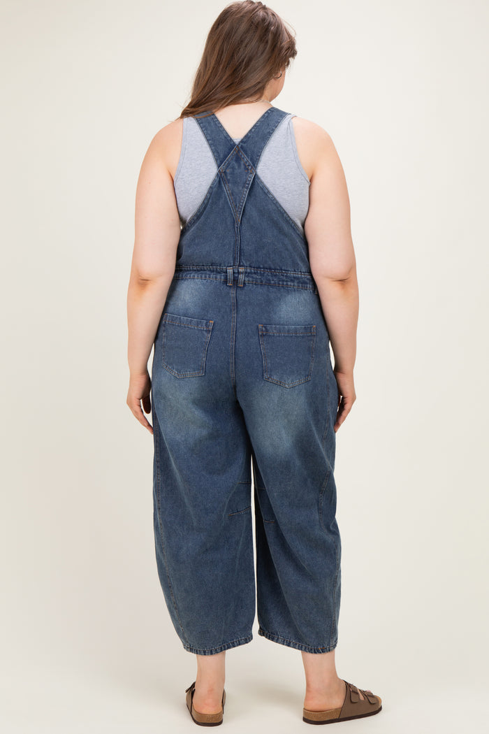 woman wearing a blue denim maternity plus overall with a relaxed fit, showing the back view and styling with a gray tank top