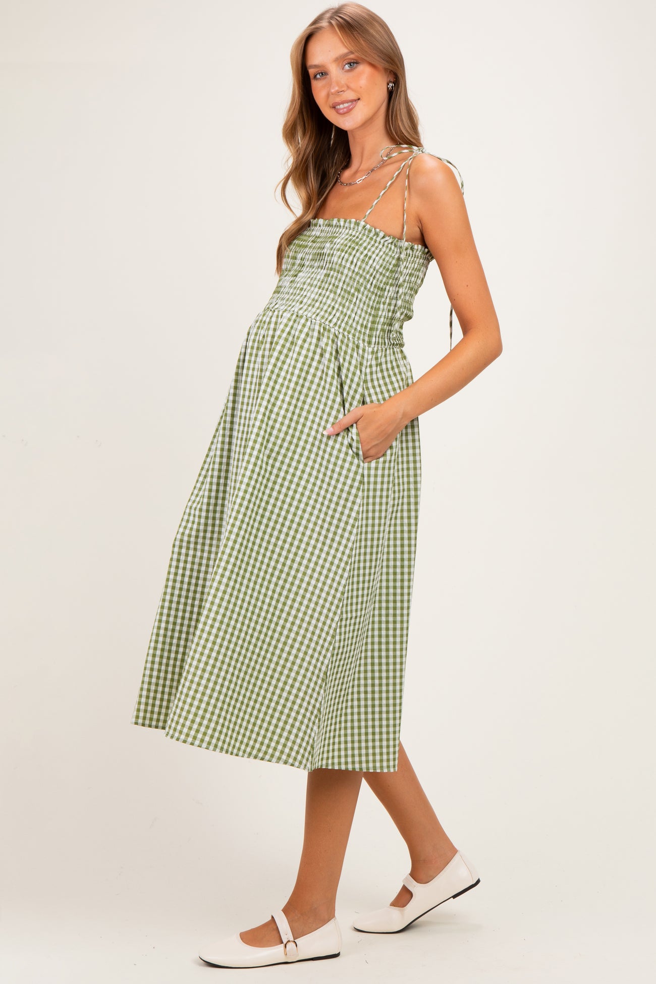 woman wearing a green and white gingham maternity dress with a smocked bodice and pockets, showing a baby bump