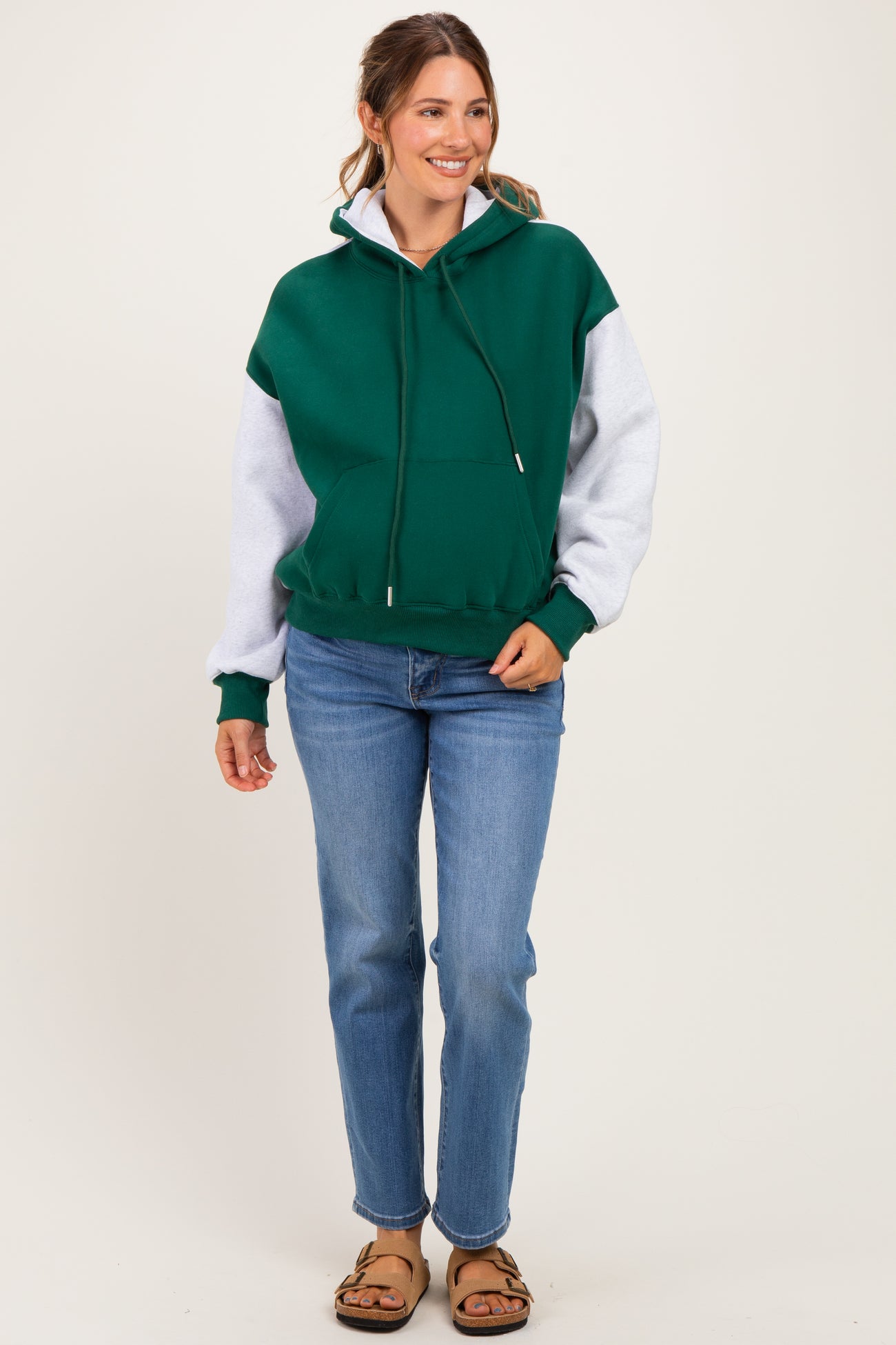 woman wearing a green and gray colorblock maternity hoodie paired with blue jeans and sandals, smiling at the camera