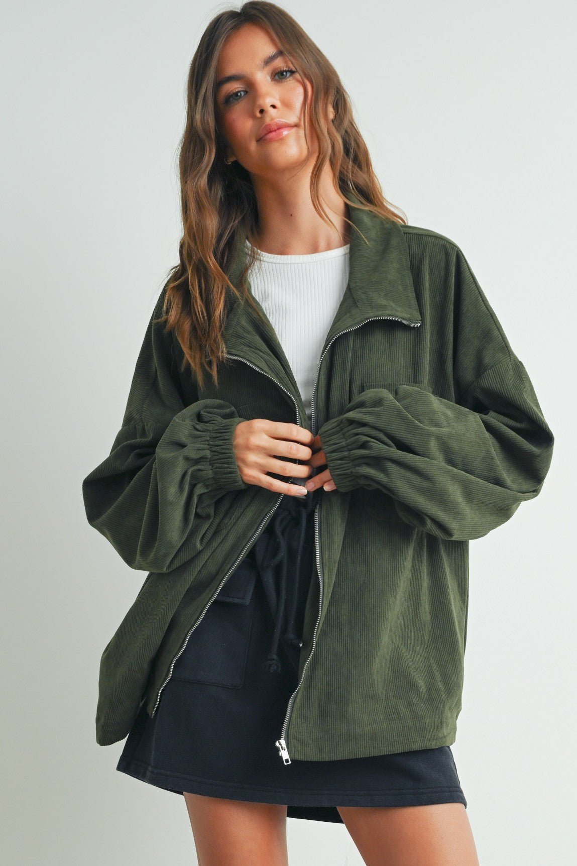 woman wearing an olive green corduroy jacket with a zip front and puffed sleeves, paired with a white top and black shorts