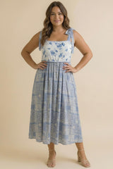 woman wearing a blue floral maxi dress with tie straps and a cinched waist, standing against a neutral background