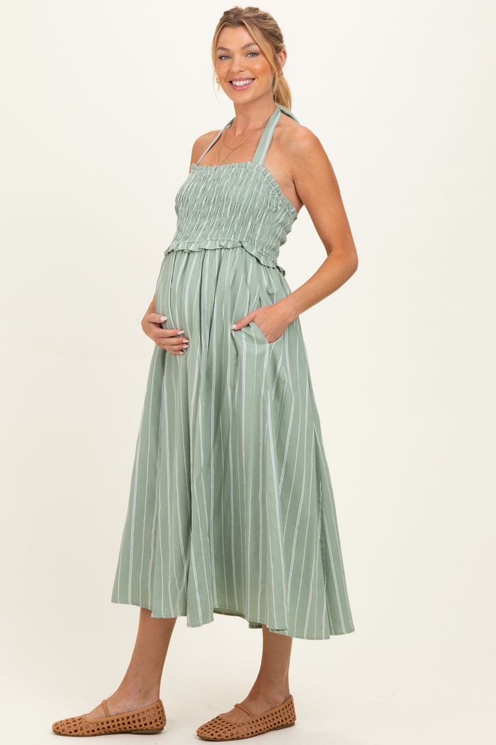 woman wearing a light green striped maternity dress with smocked bodice and pockets, holding her baby bump