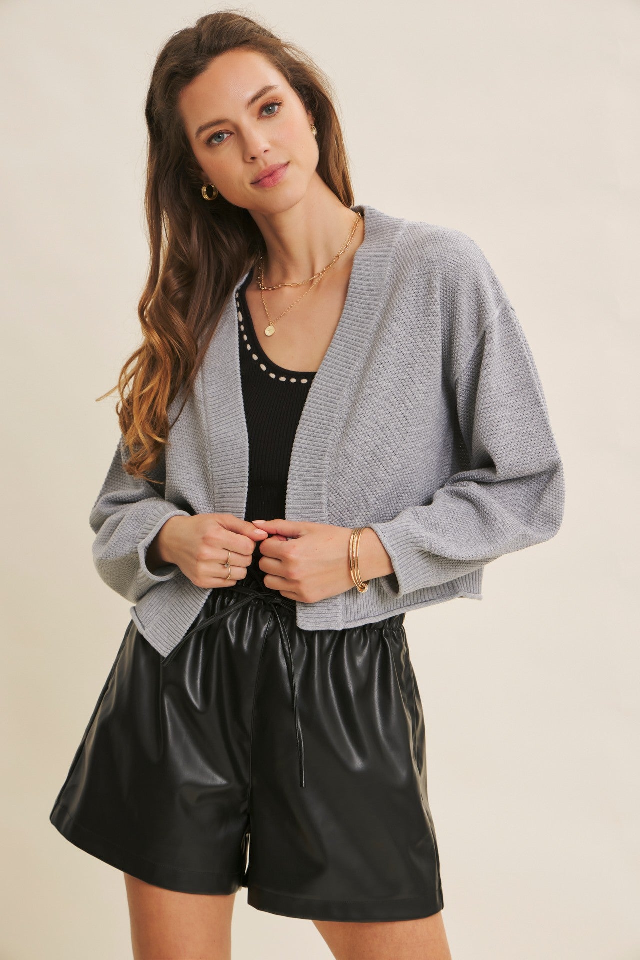 woman wearing a gray cropped cardigan over a black tank top and black faux leather shorts