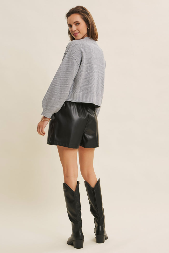 woman wearing a light gray cropped sweater and black faux leather shorts, standing with her back to the camera