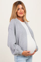 woman wearing a light gray oversized cardigan over a white top, holding her baby bump in a maternity wear style