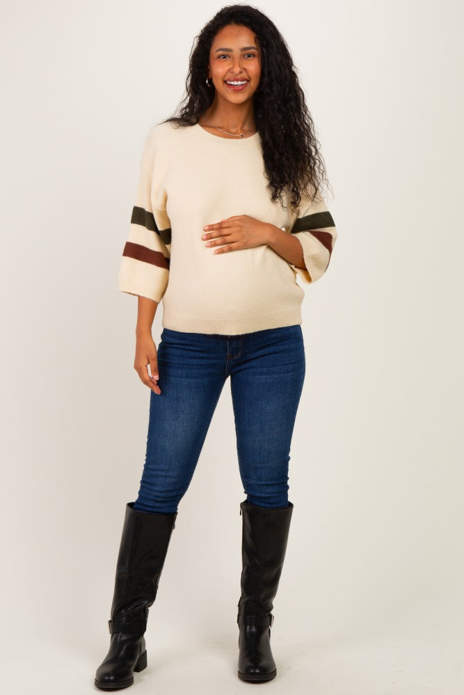 woman wearing a cream maternity sweater with striped sleeves and holding her baby bump, paired with blue jeans and black boots