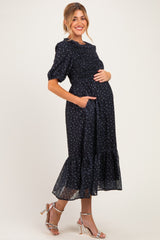 woman wearing a navy blue floral smocked maternity dress with short sleeves and a ruffled hem, holding her baby bump