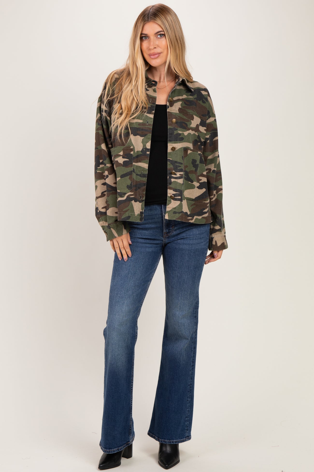 woman wearing a camouflage oversized jacket with a black top and flared jeans, standing against a neutral background