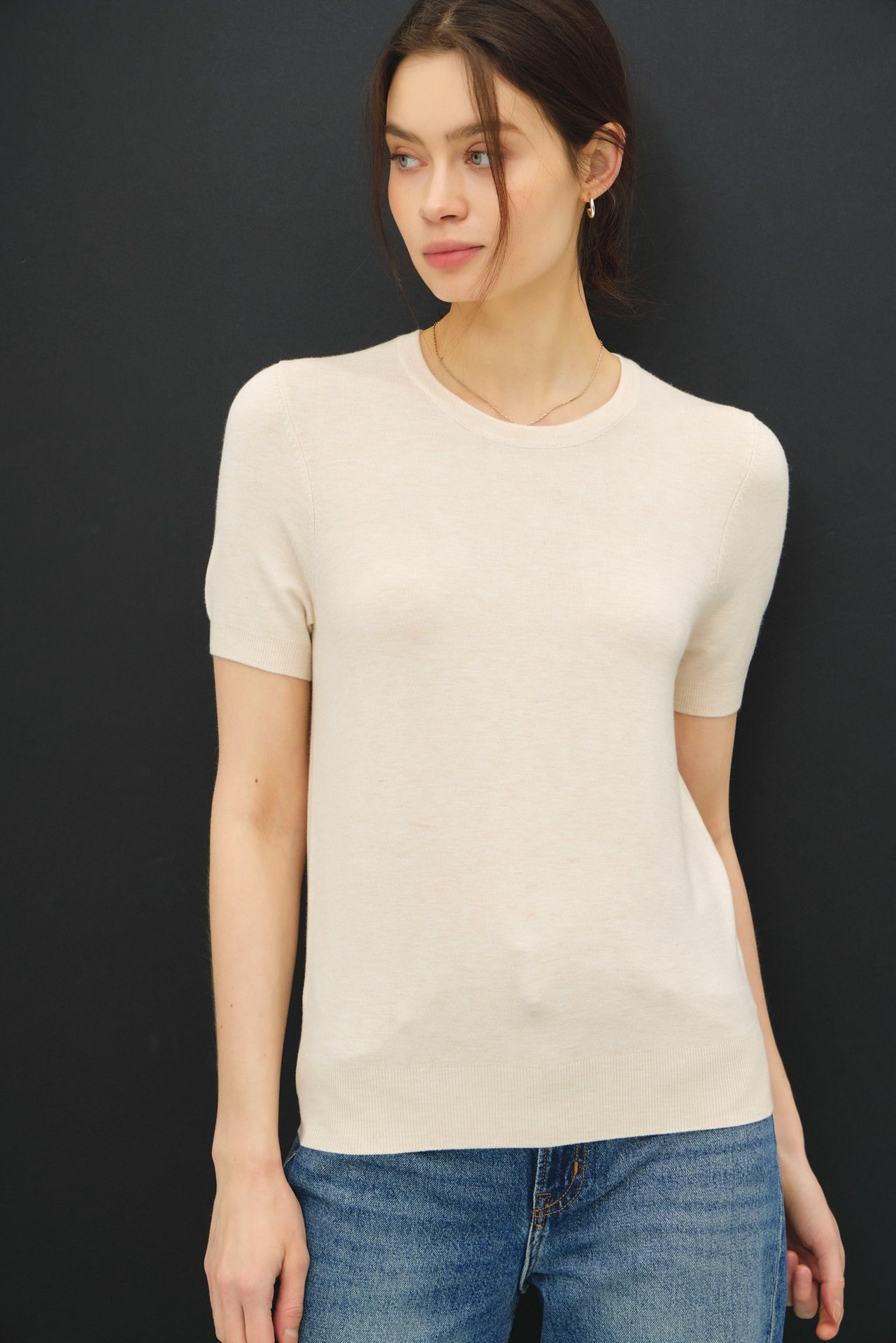 woman wearing a short sleeve beige knit top paired with light blue jeans against a dark background