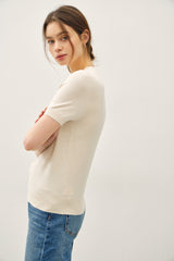 woman wearing a light beige short sleeve sweater and blue jeans, standing sideways with arms crossed