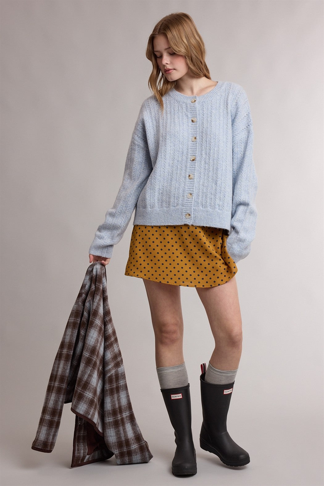 woman wearing a light blue cable-knit cardigan over a mustard polka dot mini skirt and black rain boots, holding a plaid jacket