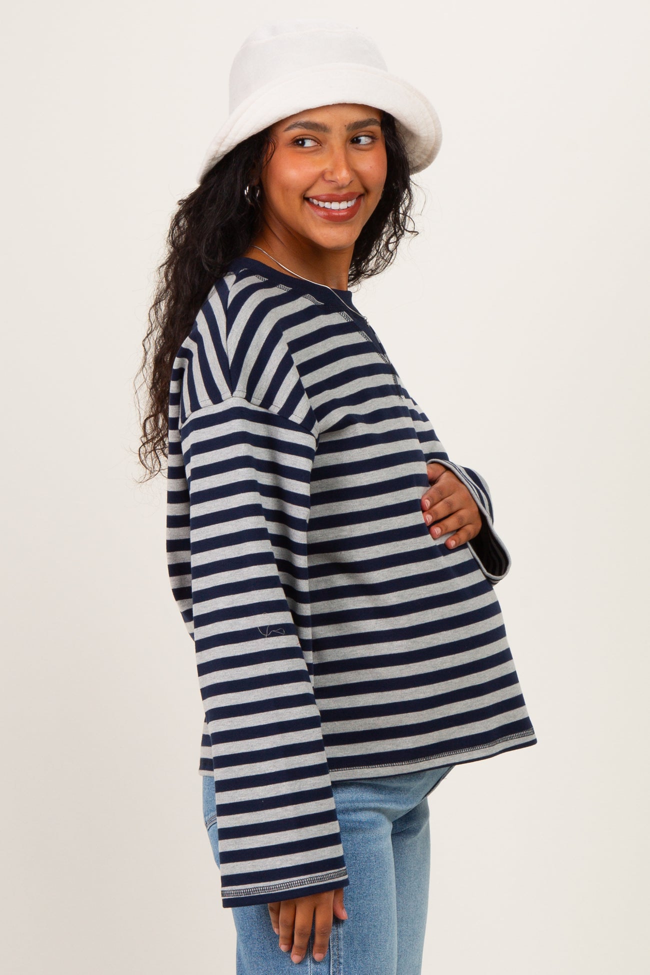 woman wearing a navy and gray striped maternity top, touching her baby bump and smiling, with a white bucket hat