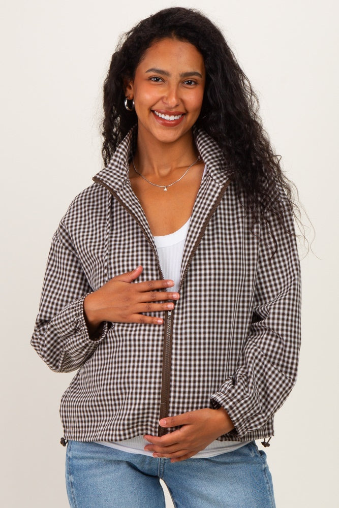 woman wearing a brown and white gingham patterned maternity jacket, gently holding her baby bump