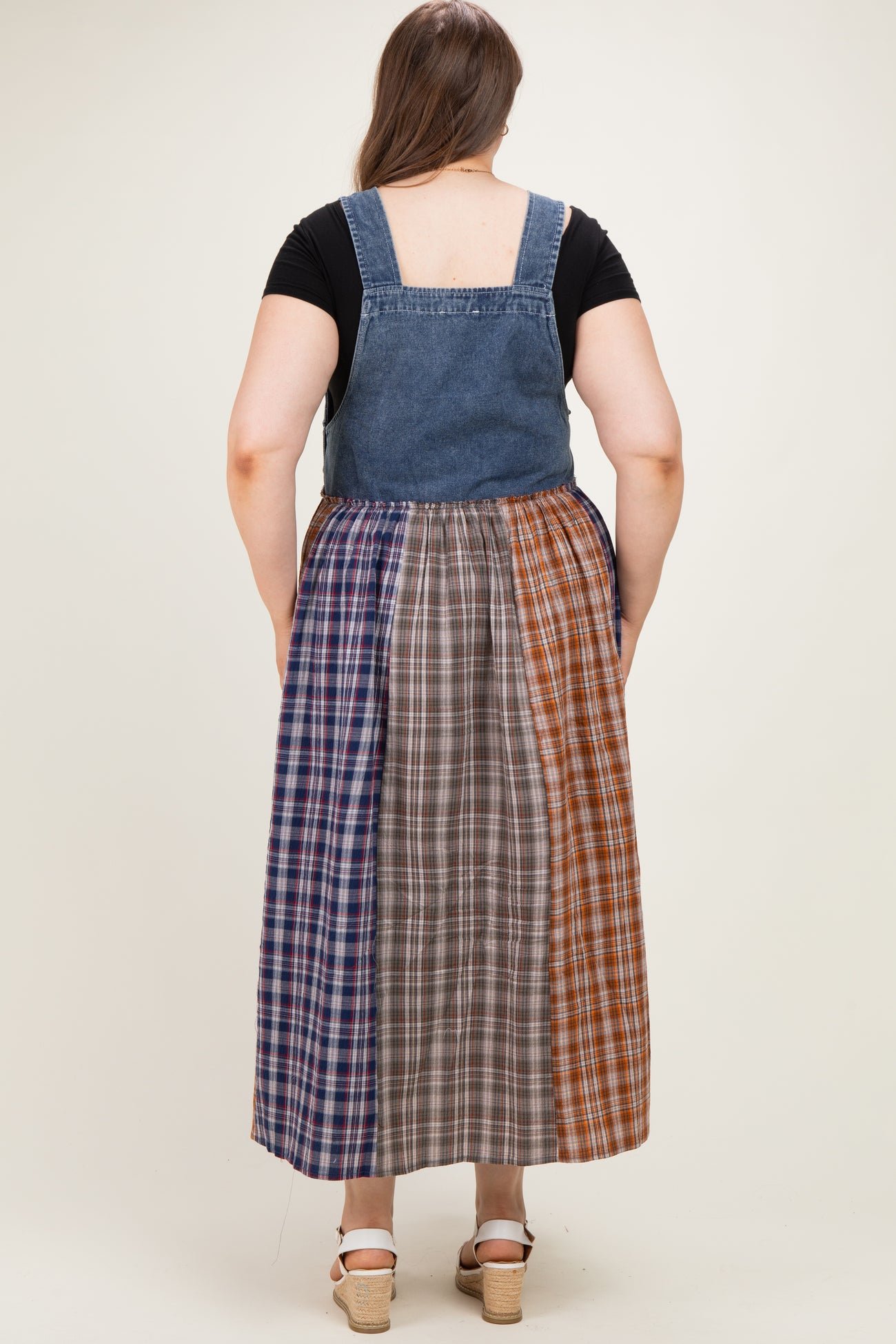 woman wearing a denim maternity plus overall dress with a multi-colored plaid skirt in a full length style from the back