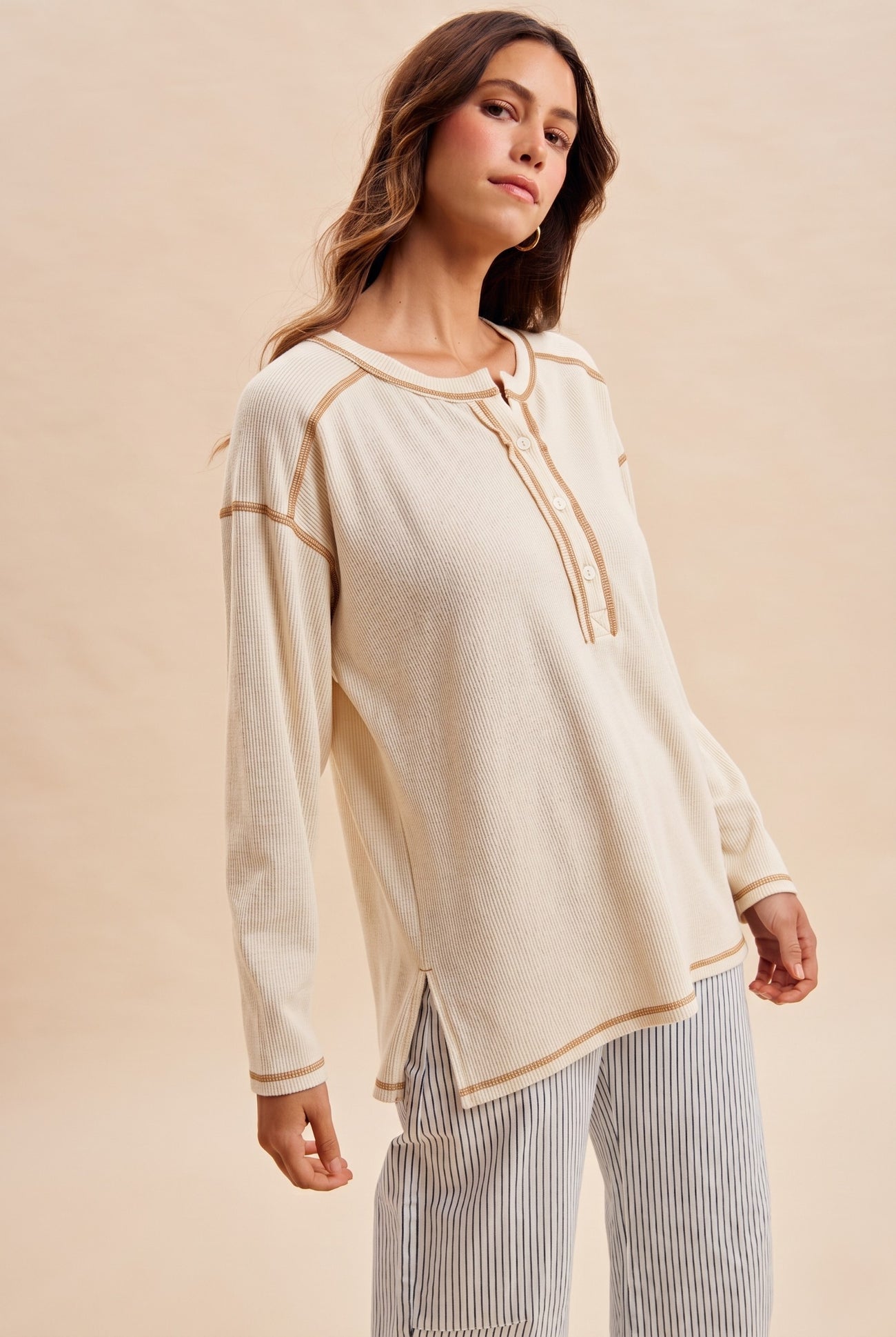 woman wearing a beige long sleeve henley top with button details and contrast stitching, styled with striped pants