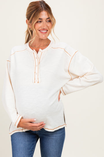woman wearing a cream maternity top with button details and contrasting stitching, holding her baby bump