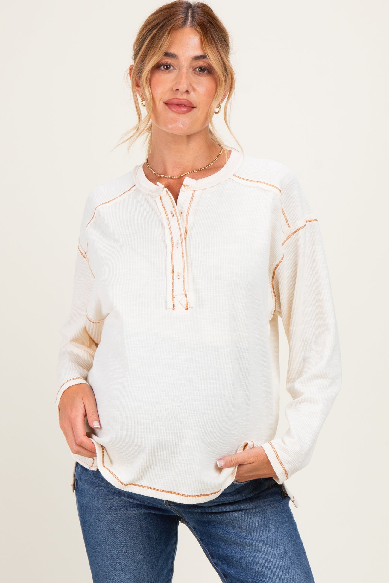 woman wearing a cream maternity top with a henley neckline and orange stitching, showing her baby bump