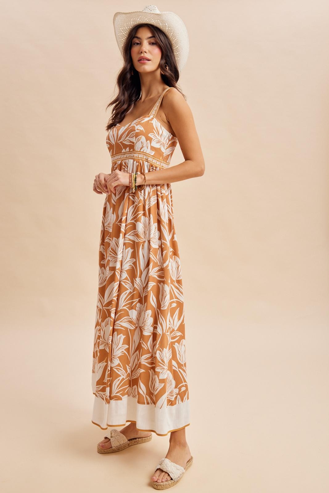 woman wearing a brown floral maxi dress with spaghetti straps and a white hem, accessorized with a wide-brimmed hat