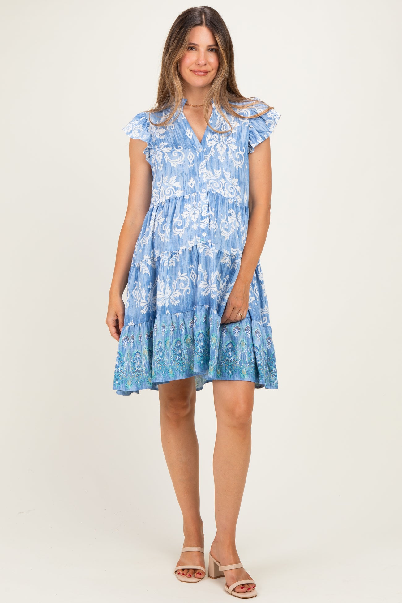 woman wearing a blue floral print maternity dress with ruffled sleeves and a tiered skirt, standing and smiling