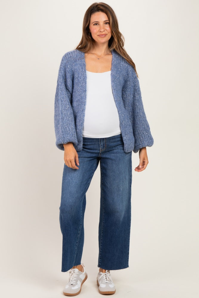 woman wearing a light blue fuzzy maternity cardigan over a white top and blue wide-leg jeans, showing her baby bump