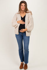 woman wearing a beige knitted maternity cardigan and black top, holding her baby bump with a smile