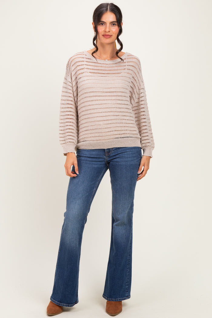 woman wearing a light brown open knit sweater and blue flared jeans, standing with a neutral background