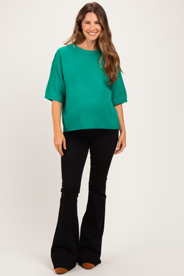 woman wearing a green oversized maternity sweater with short sleeves and black flared pants, showing her baby bump