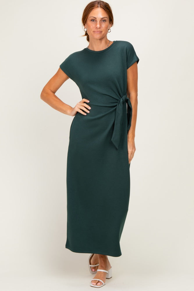 woman wearing a dark green maxi dress with a side tie detail and short sleeves, standing on a neutral background