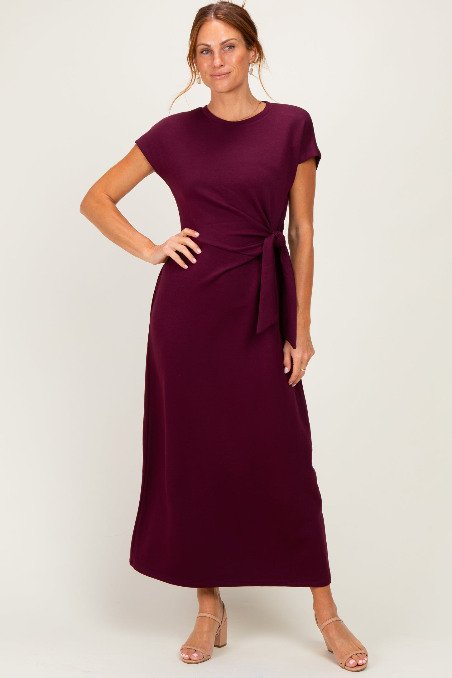 woman wearing a burgundy maxi dress with short sleeves and a knot detail at the waist, posing with one hand on her hip