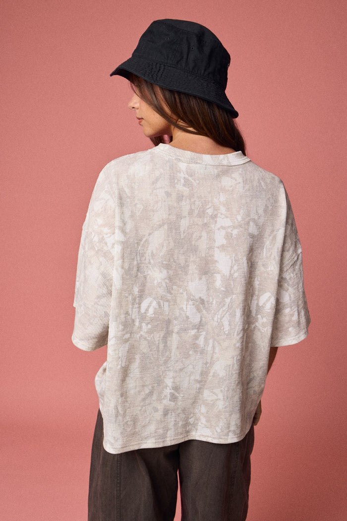 woman wearing an oversized beige tie-dye shirt and a black bucket hat, viewed from the back against a pink background