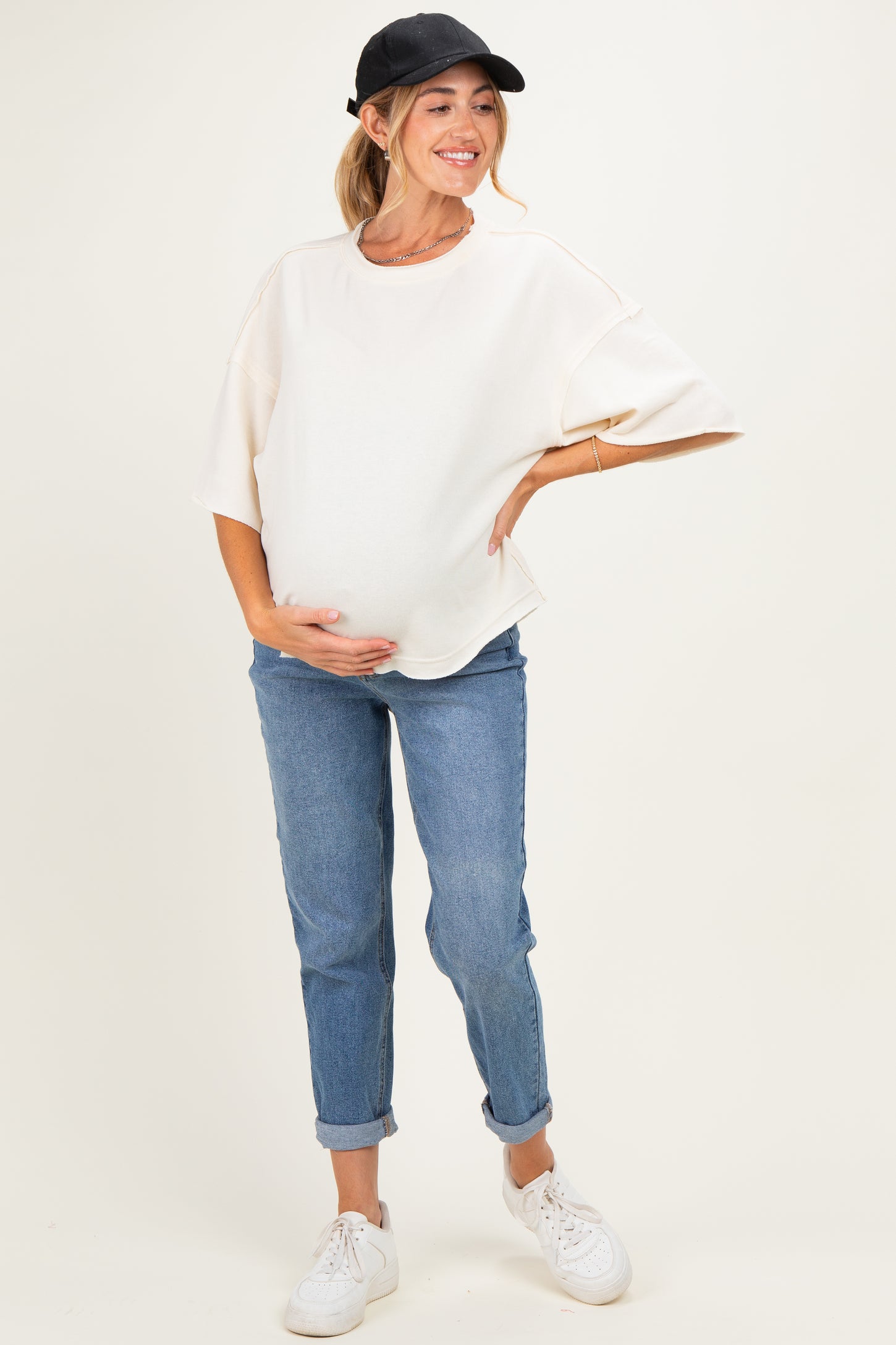 woman wearing a cream oversized maternity top and blue jeans, holding her baby bump while smiling.