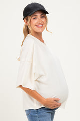 woman wearing a cream oversized maternity top and blue jeans, gently cradling her baby bump