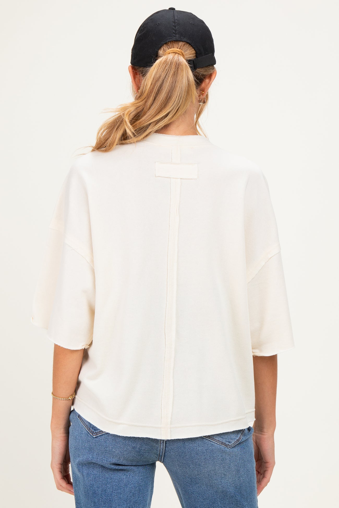 woman wearing a cream-colored maternity top with a relaxed fit and back seam detail, styled with a black cap