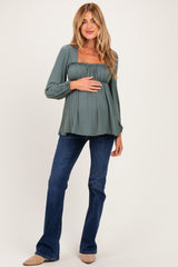 woman wearing a sage green long sleeve maternity top with smocked detailing and a visible baby bump, paired with flared jeans