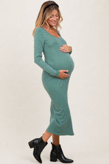 woman wearing a green long sleeve maternity dress, cradling her baby bump, and standing in a side profile