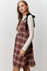 woman wearing a maroon plaid pinafore dress over a white long sleeve top with black ribbon details